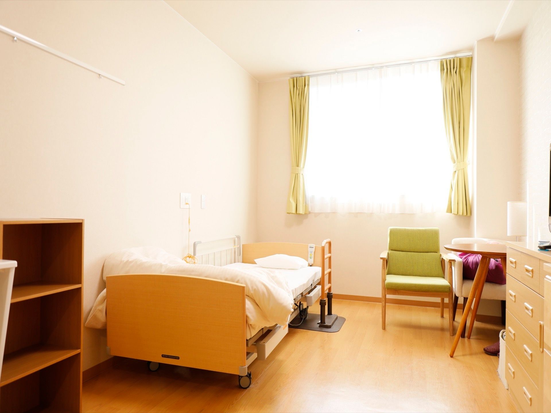 ワンルームの部屋に介護ベッドやテーブル、椅子などの家具が置かれている。壁にはナースコールが設置されている。