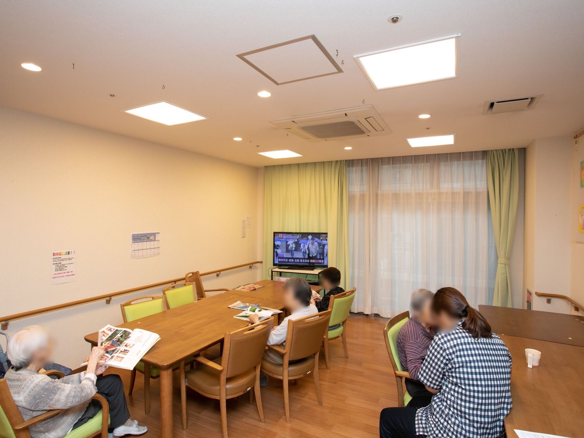 ダイニングテーブルがいくつも並んでいる。入居者様がテレビを鑑賞しながら、自由に過ごしている。大きな窓にはカーテンが掛けられている。