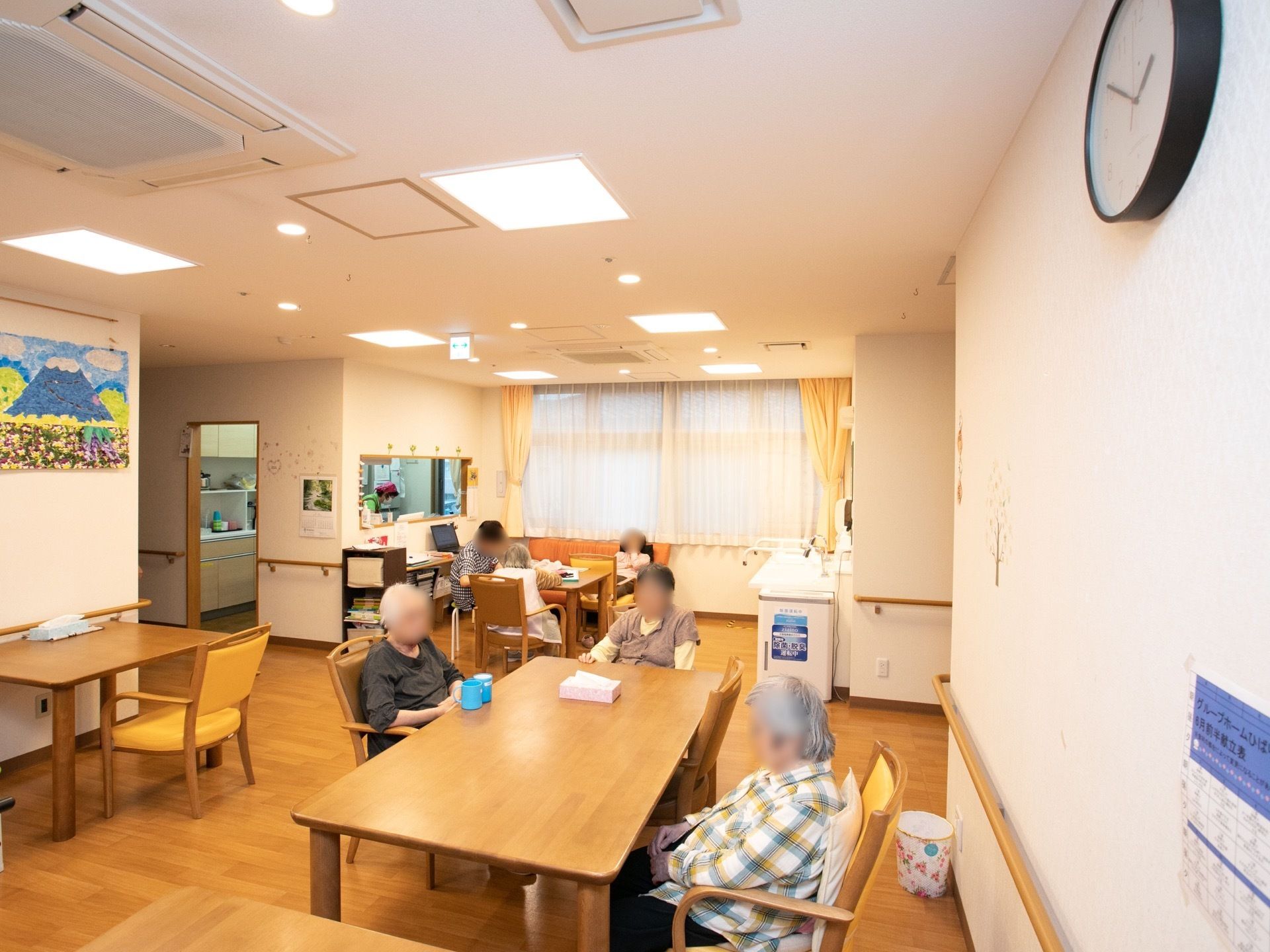 キッチンが併設されている。入居者様が席につき、くつろいでいる。壁には入居者様が製作した作品がレイアウトされている。