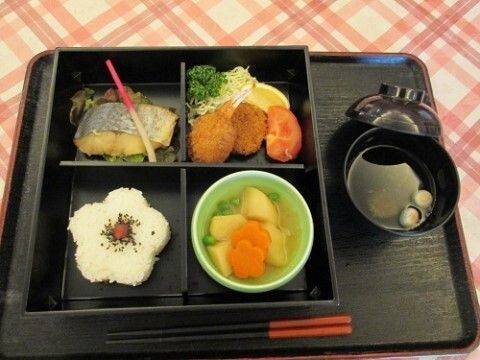 黒いお盆の上に食事が配膳されている。ご飯は花の形に模られており、中心には梅干しと胡麻が盛りつけられている。魚やコロッケもある。