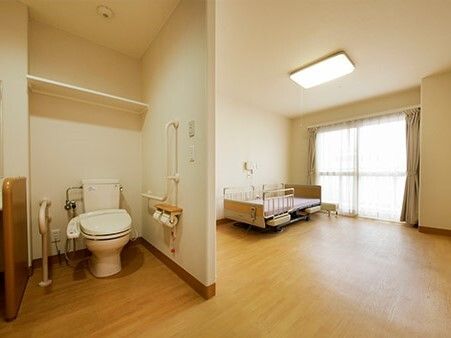 レースカーテンとカーテンが付いた大きな窓があるフローリングの部屋には寝具付きのベッドやトイレがあり、緊急通報装置も付いている。