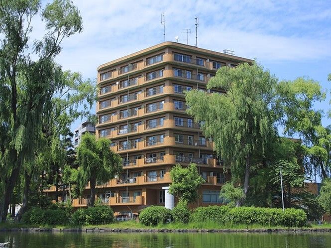 オレンジ系の外壁のマンションのような大きな建物。近くには川のような場所があり、緑も多く、自然に囲まれている。
