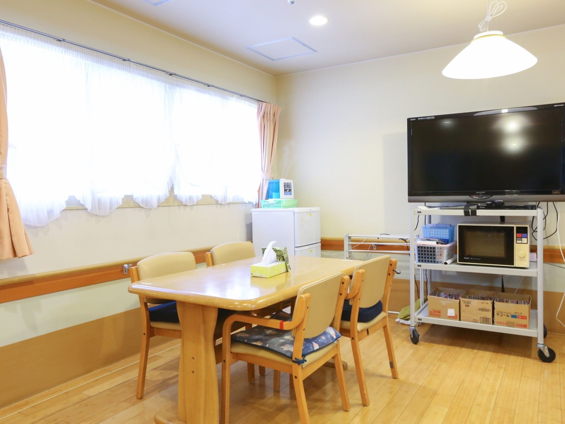床がフローリングの食堂には肘掛け付きの椅子が４脚とテーブルが１台置かれている。大型テレビや冷蔵庫なども設置されている。