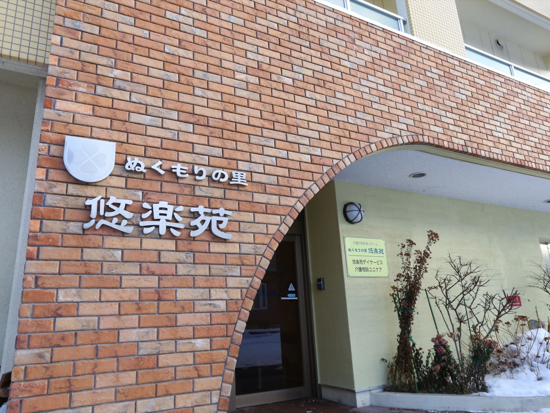 施設の外壁はレンガ造りになっており、施設の名前のプレートが取り付けられている。玄関の脇には様々な植物が植えられた花壇がある。