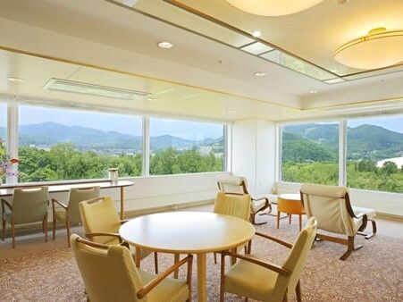 窓のそばには一人用の椅子が点々と置かれていて、部屋の中央には丸いテーブルセットが置かれ、窓の外には緑が見える。