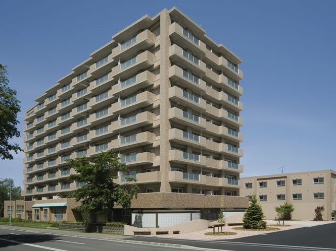 各フロアにベランダが付いた大きな建物はベージュ系でまとめられ、周りには緑がたくさん植えられ、奥にもベージュの建物がある。