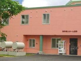 ピンクの外壁の２階建ての建物の外壁にはホーム名が書かれ、ドアの横には自動販売機が設置されている。