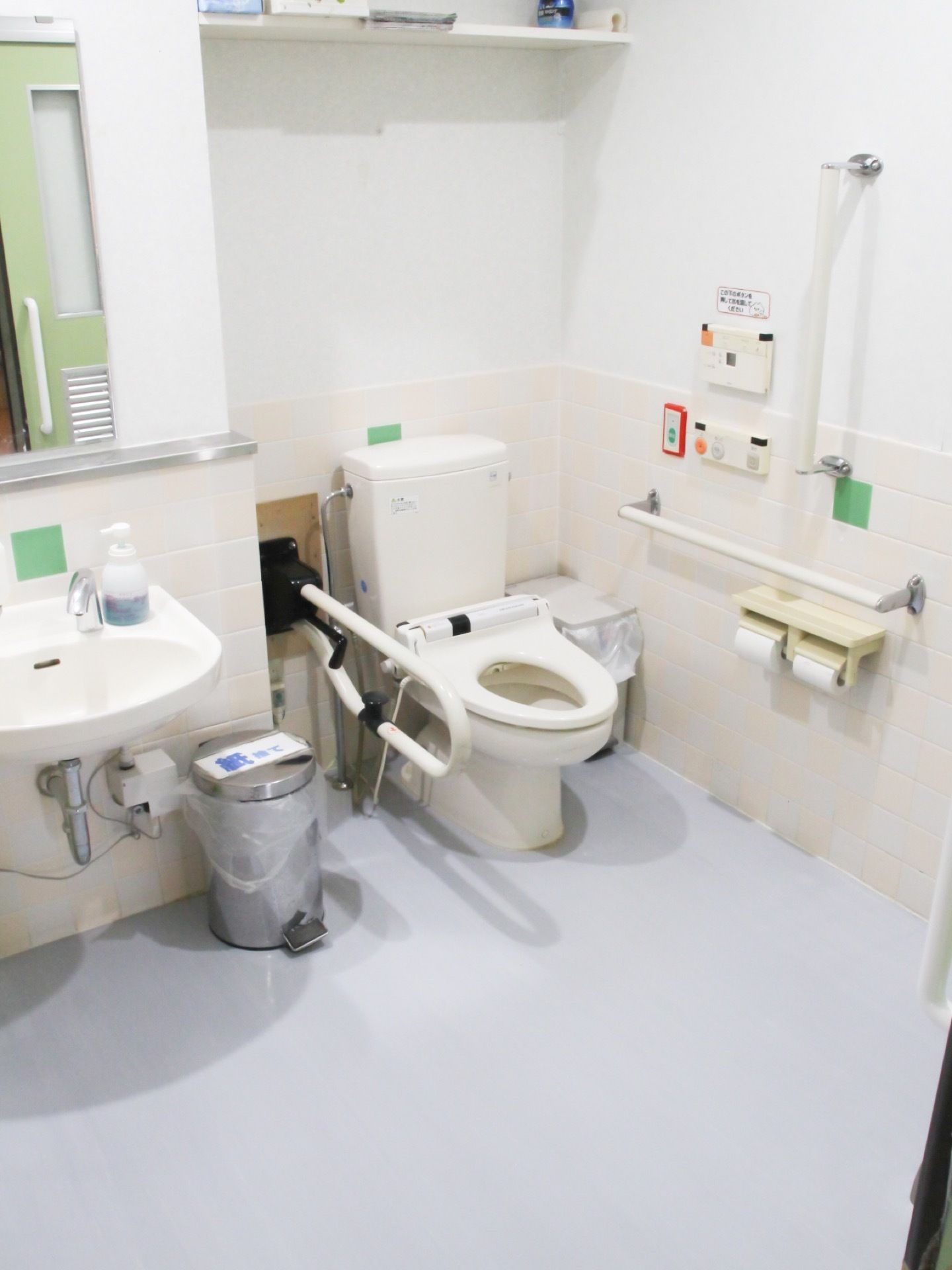 タンク付きの白い洋式トイレの横に洗面台が設置されている。洗面台の前の壁に鏡があり、後ろの扉が映っている。