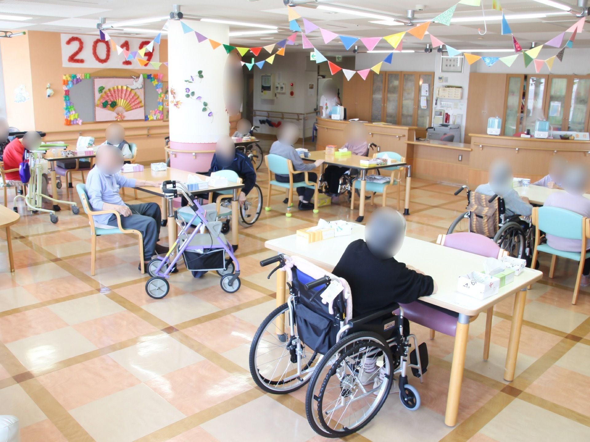 部屋の中に円柱の柱が立っている。たくさんのダイニングテーブルや椅子が置いてあり、それぞれに入居者様が腰を掛けている。