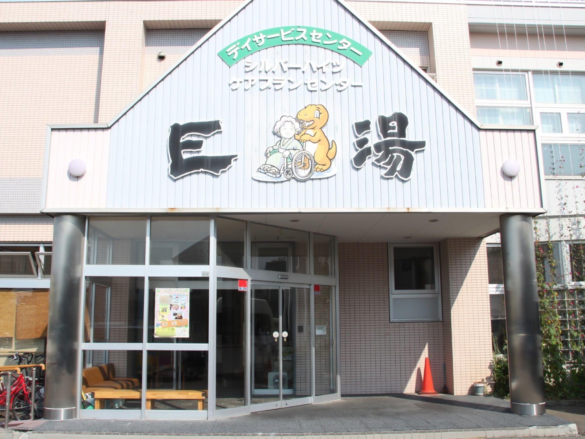 建物の入口の屋根に施設の名称と老人が乗った車椅子を押す恐竜の絵が表示されている。赤い三角コーンが置いてある。
