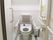 壁にタイルが貼られた個室のトイレがある。トイレの扉が開かれ、背もたれと手すりが設置された洋式便器が見えている。