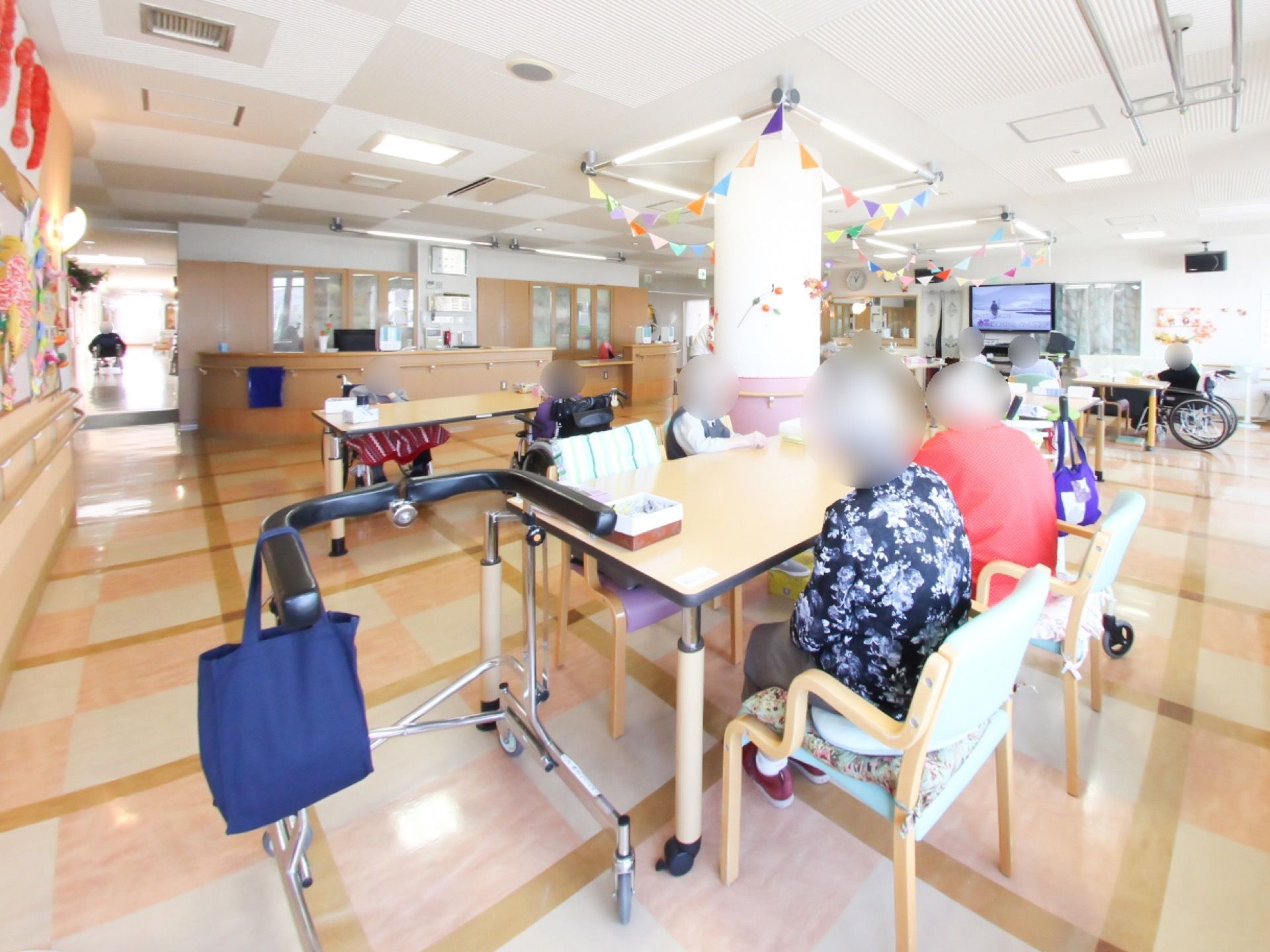 テーブルの横に歩行器が置いてあり、数名の入居者様が腰を掛けてテレビを見ている。円柱型の柱に飾り付けがされている。