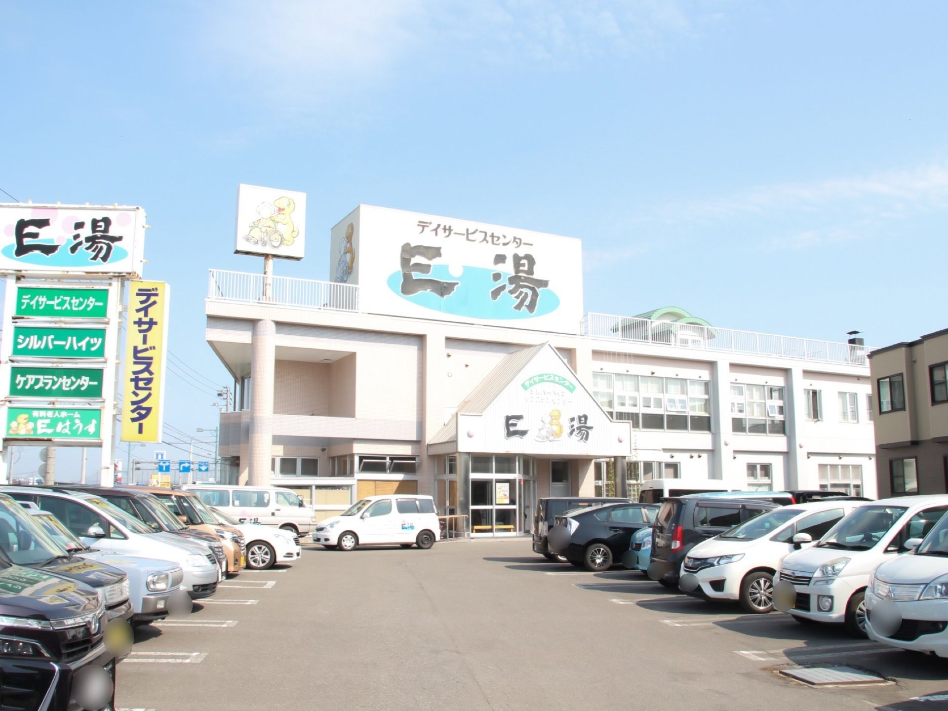 ２階建ての建物の前に白線で区切られた駐車場があり、たくさんの車が停められている。施設名が表示された大きな看板が立てられている。
