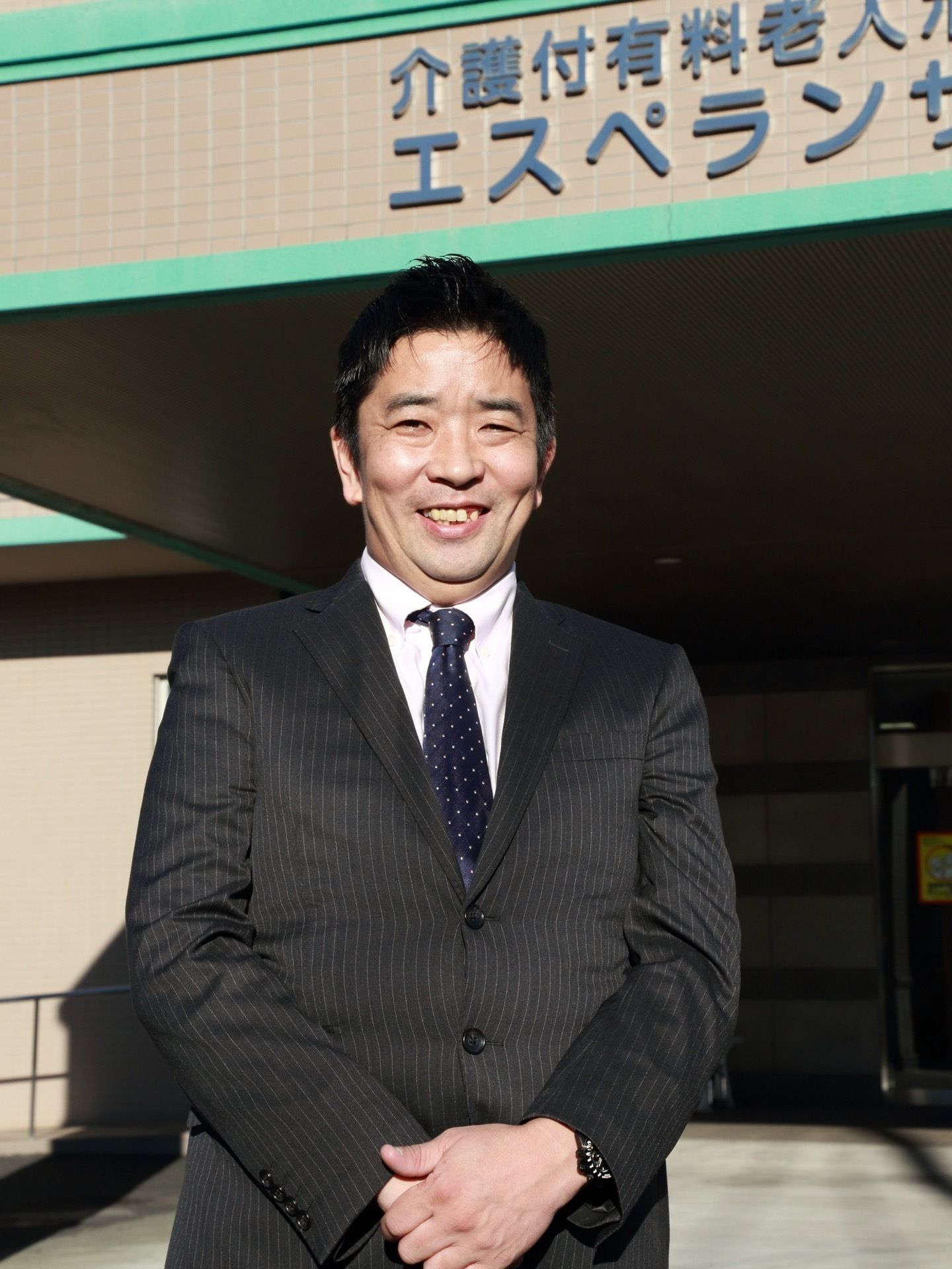 建物の入り口前で笑顔を浮かべる、両手を前で重ね合わせて立つスーツ姿の男性