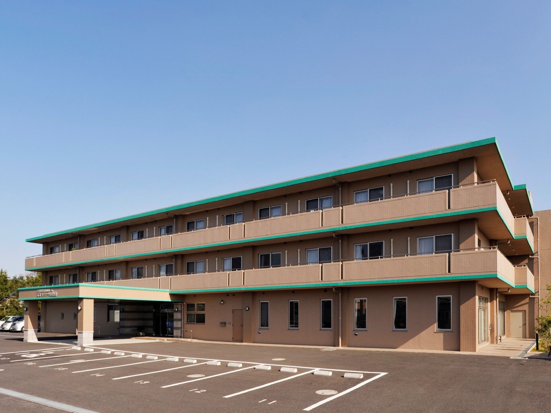 明るい茶色の外壁がマンションのような施設。玄関は入り口が突き出て、屋根になっているので車から降りて濡れずに入れる。