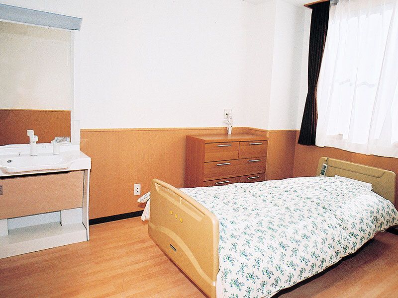 レースカーテンとカーテンが付いた窓があるフローリングの部屋には寝具付きのベッドがあり、車いす対応の洗面所もある。
