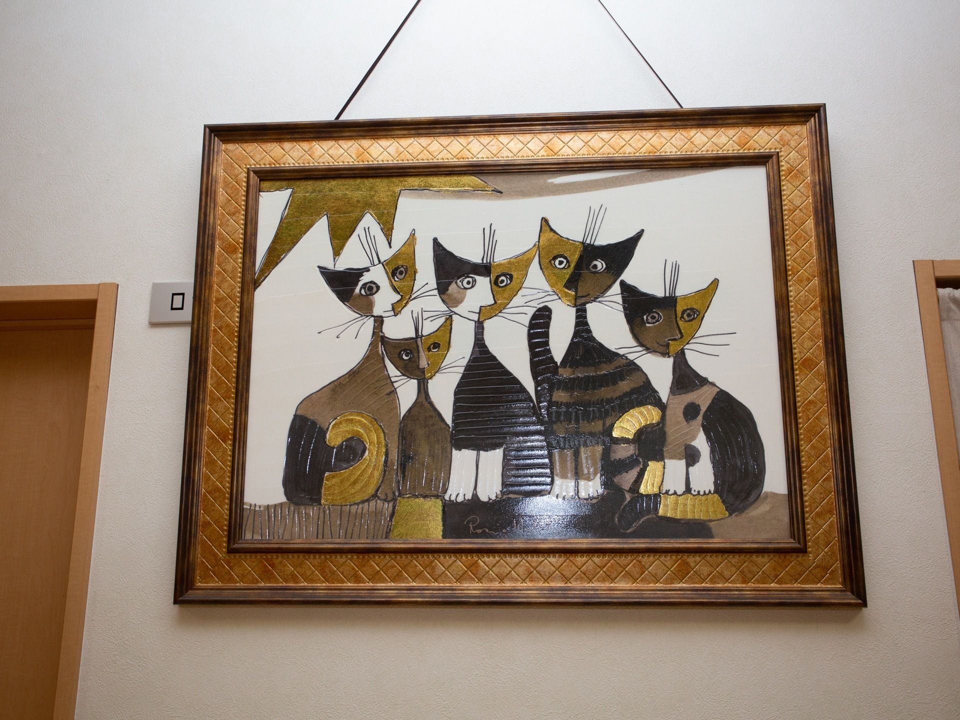 白い廊下の壁に飾られた絵画である。大きな茶色い額縁のなかに、猫が５匹描かれたイラストが入れられている。