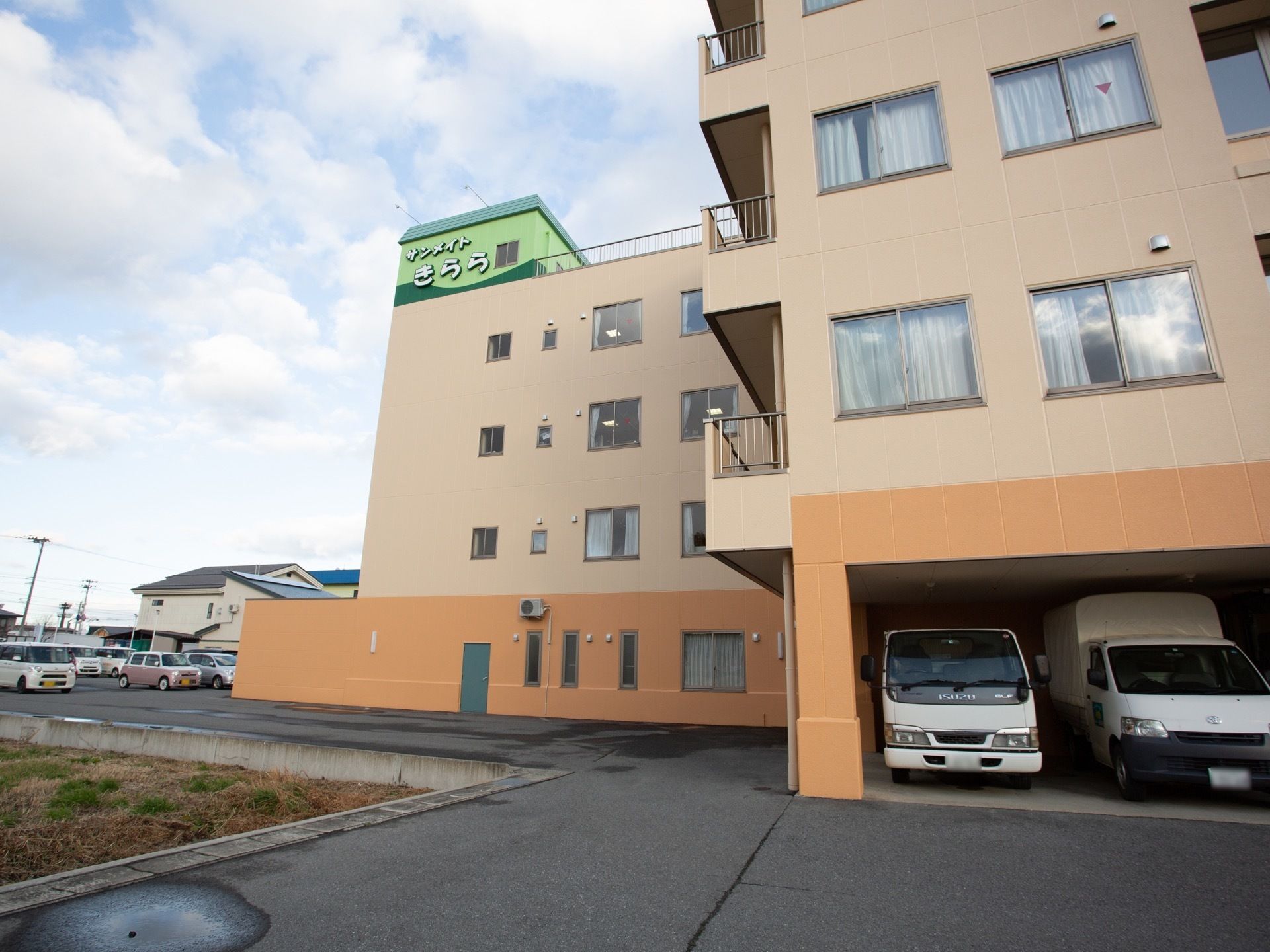 １階部分はオレンジ、２階より上はベージュに塗られた建物である。１階部分には駐車場があり、トラックなどが停まっている。