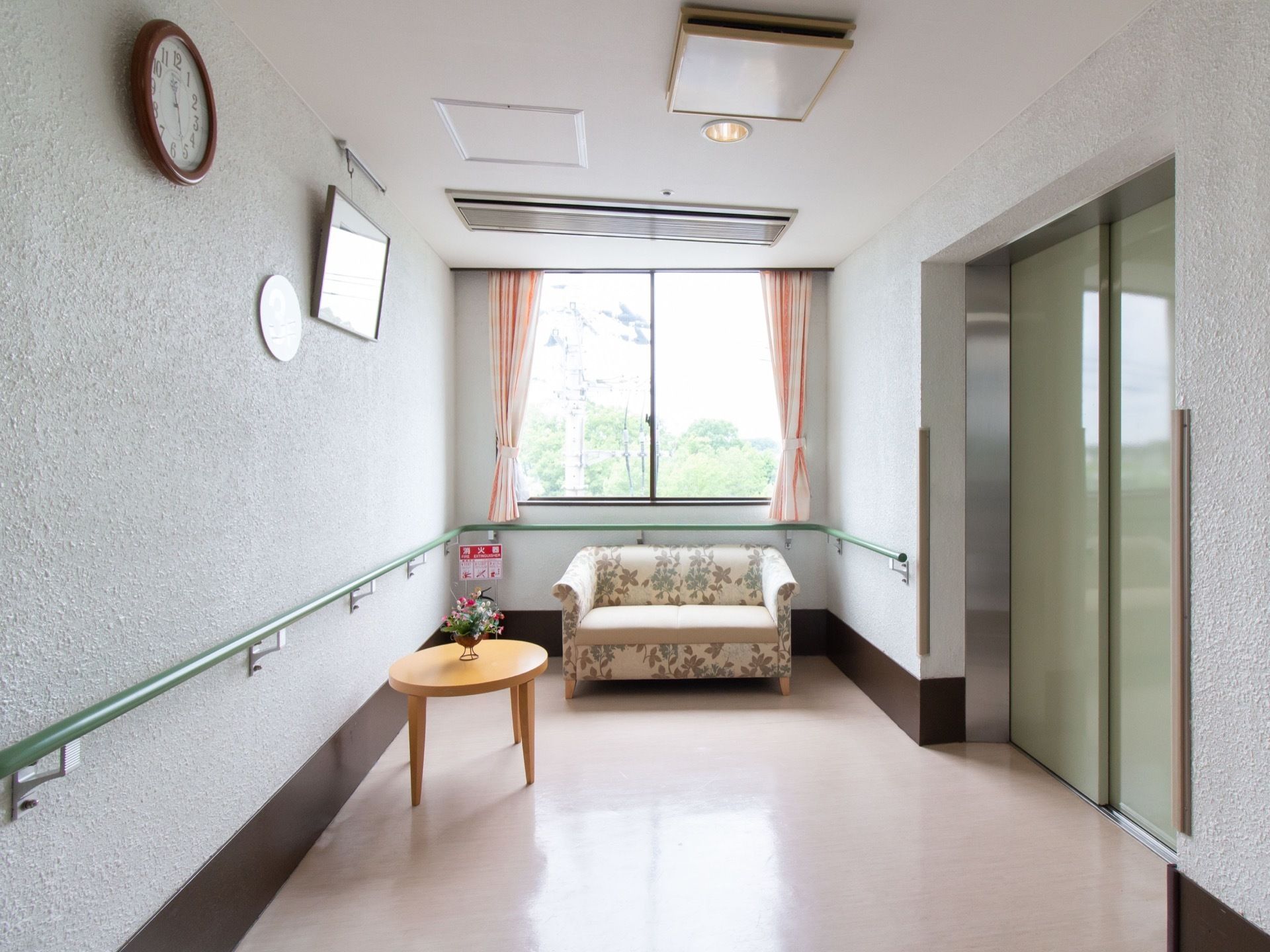 通路の突き当たりに窓があり、その手前に花柄のソファや円形テーブルが置かれている。右側に黄緑色のエレベーターの扉が見える。