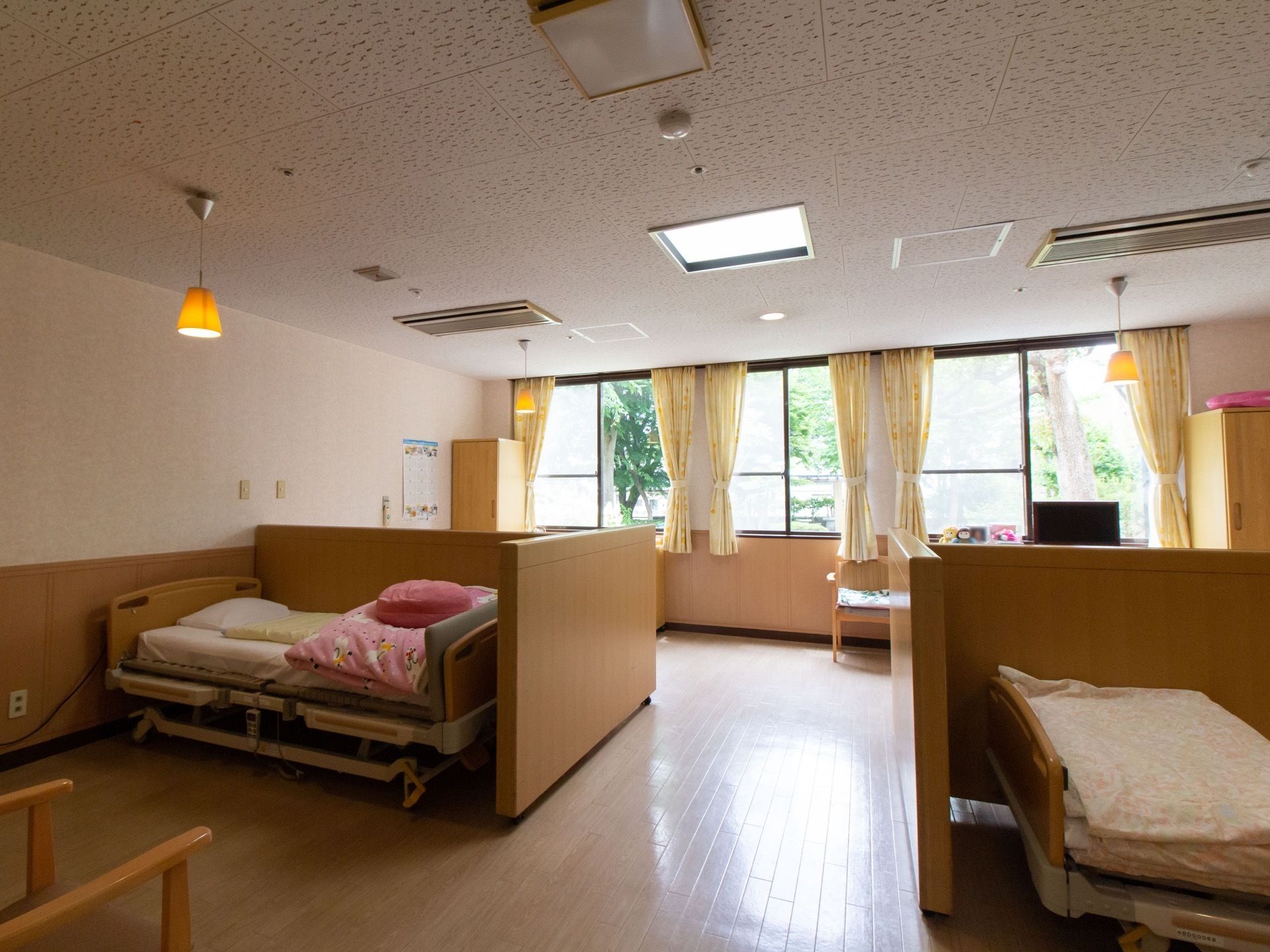 広い室内にＴ字型の仕切りが施されており、その手前にベッドが置かれている。窓の外には木々が茂っている。