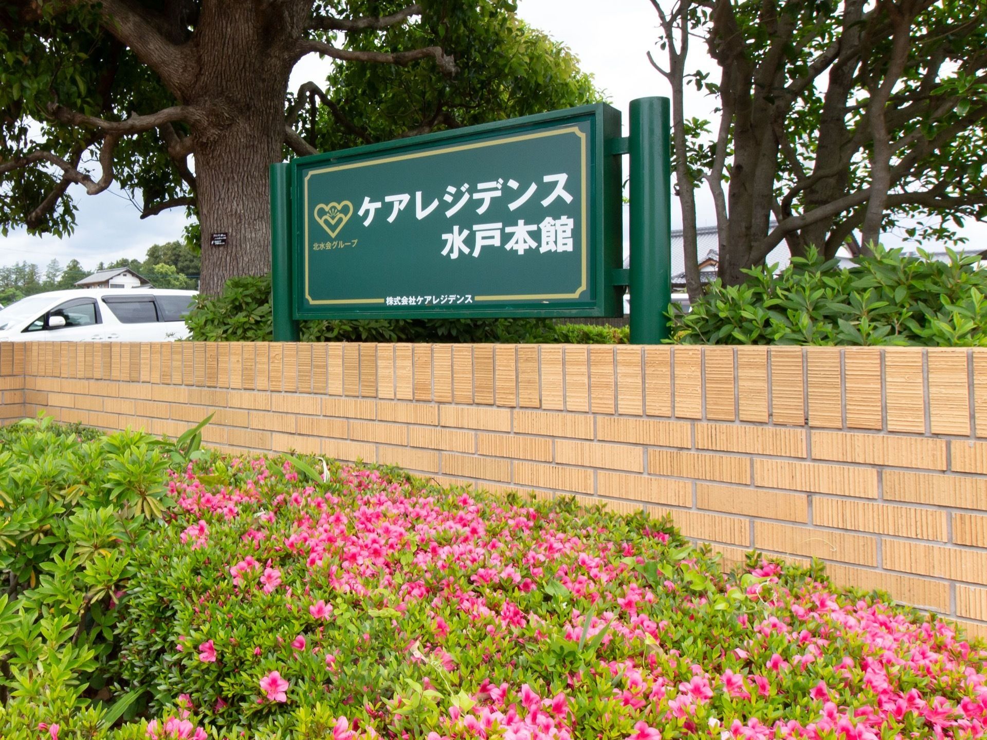 レンガ調の外壁の上方に、施設名称が明記された緑色の看板がある。外壁の外側と内側に手入れの行き届いた木々が茂っている。