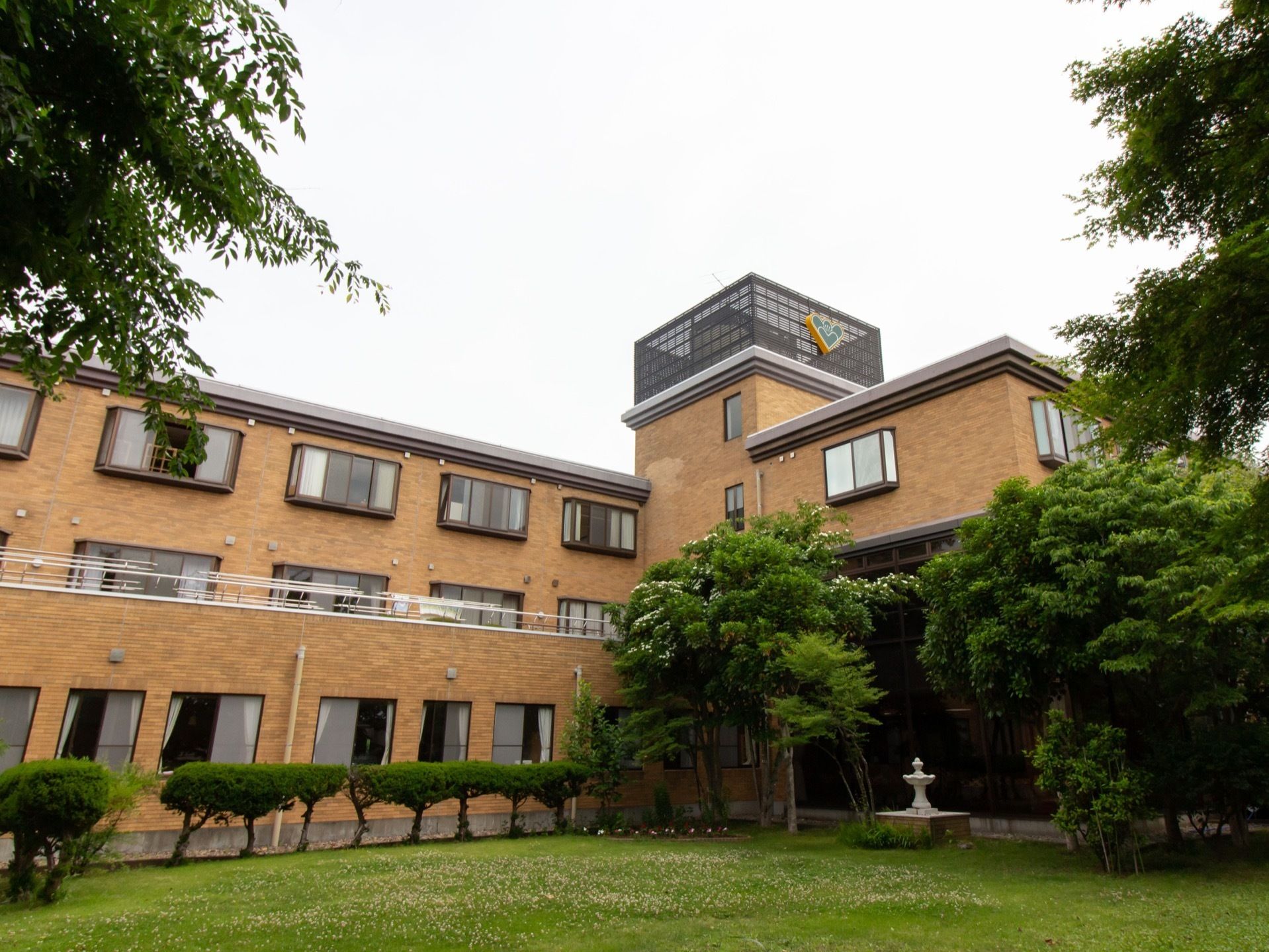 Ｌ字型の施設があり、その前のスペースに丈の短い草が生い茂っている。建物の周囲には、さまざまな種類の木が植えられている。