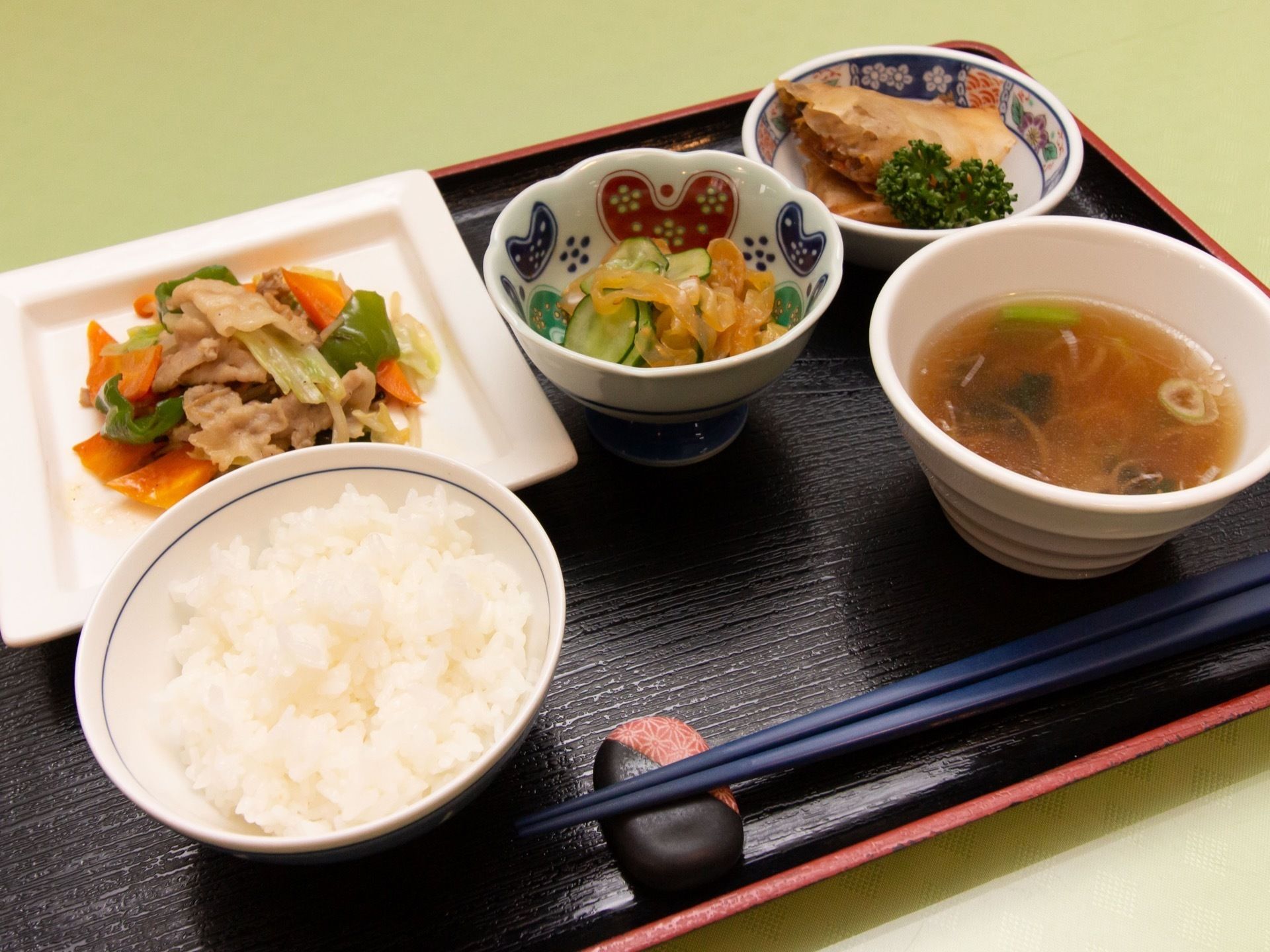 白いごはんとスープの奧に、肉野菜炒め、きゅうりの和え物、春巻きが並んでいる。さまざまな種類の野菜の彩りや器の絵柄が美しい。