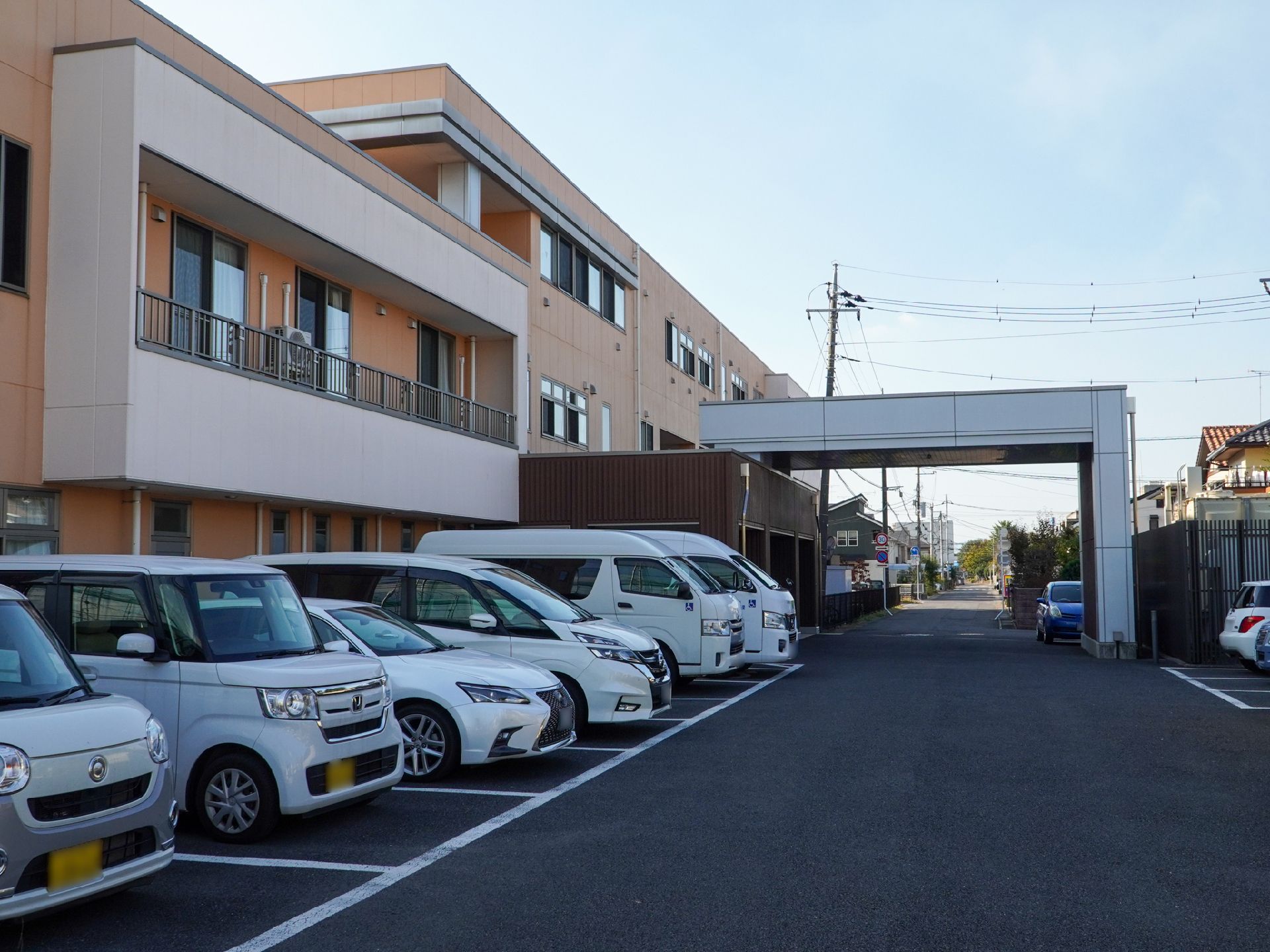 ブルーのエプロンを付けた若い女性スタッフに付き添われて屋外で過ごしている車いすの高齢女性。ニット帽をかぶっている。