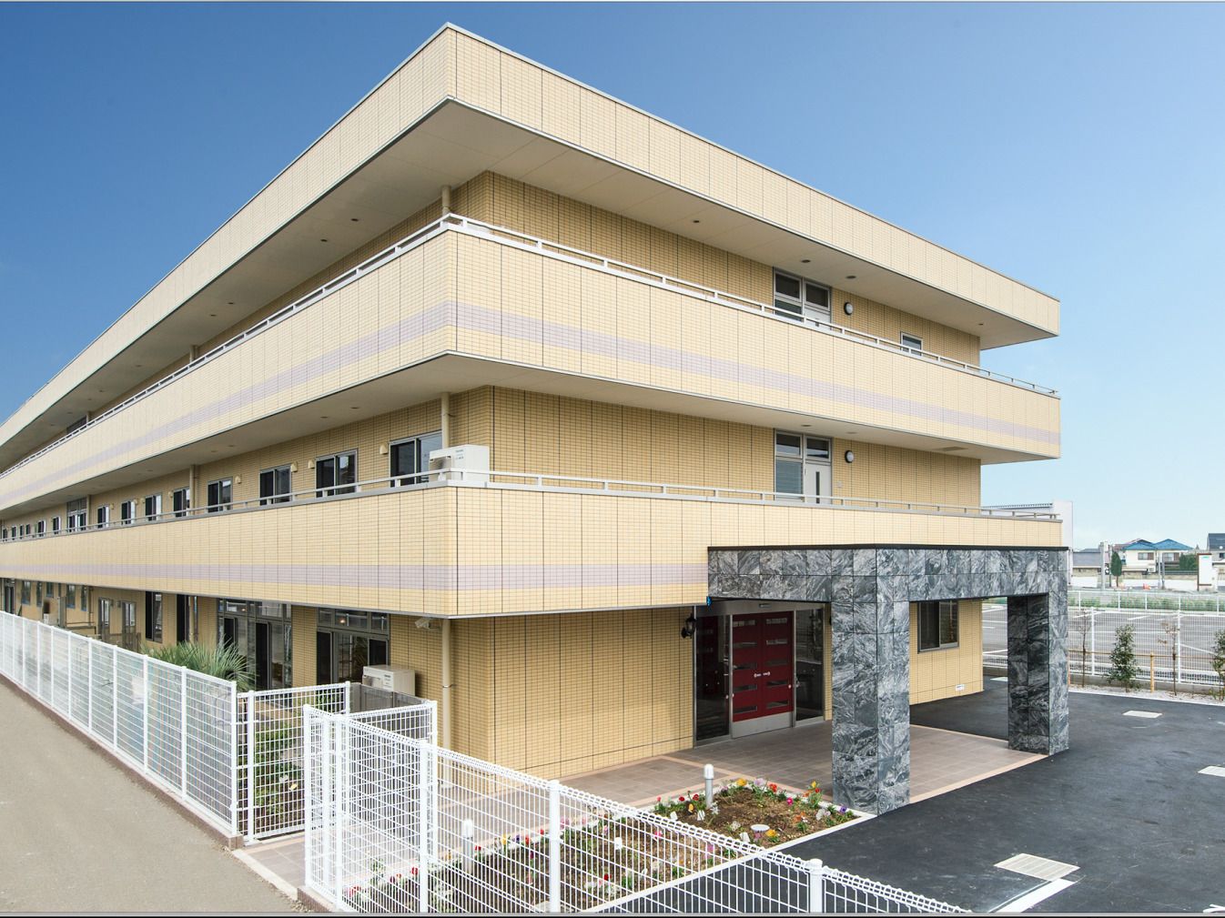 車寄せが付いている玄関がある3階建ての建物はベージュ系の外壁で、玄関前には花壇もある。