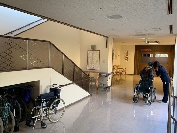 廊下でひそひそ話をしている女性入居者と女性スタッフ。入居者は車いすに寄りかかり立っている。