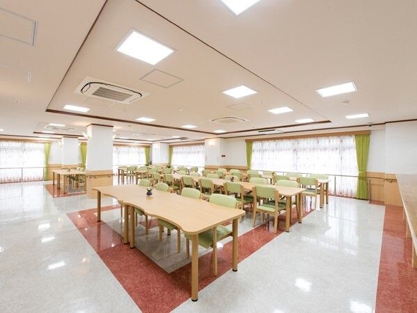 グリーンの椅子がセットされた木製のテーブルが複数並んだ広い食堂。大きな窓がたくさんある。