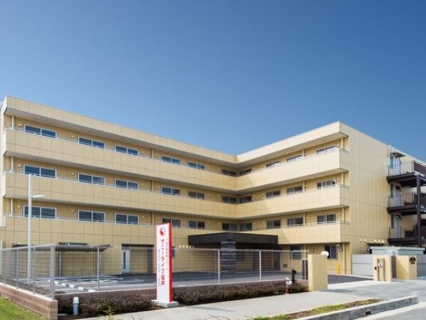エル字型のベージュの外壁の建物は4階建てで、敷地内には駐車場があり、館銘板もある。