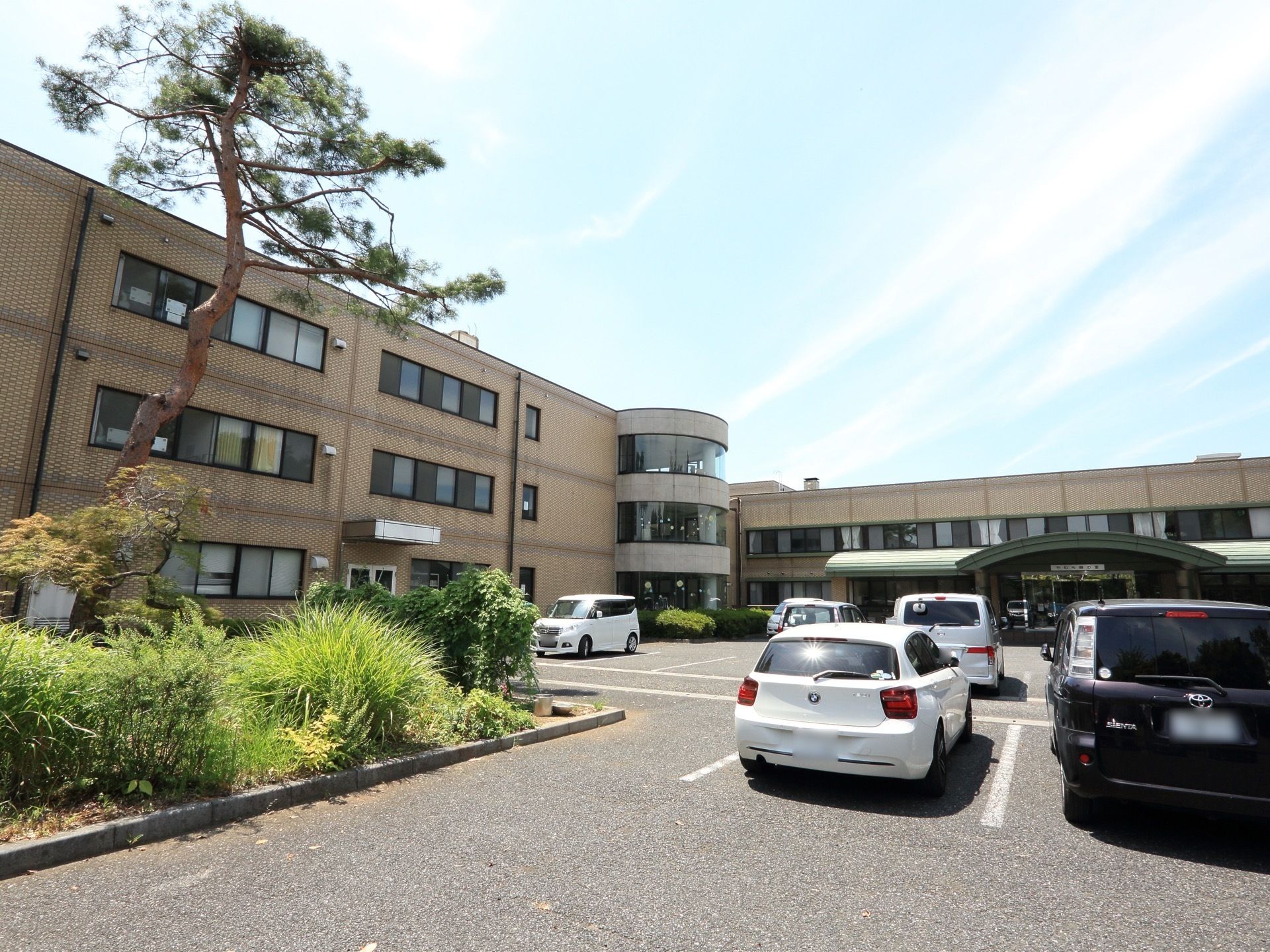 建物の前には広い駐車場があり、車がたくさん停まっている。まわりには花壇があり、植物が植えられている。