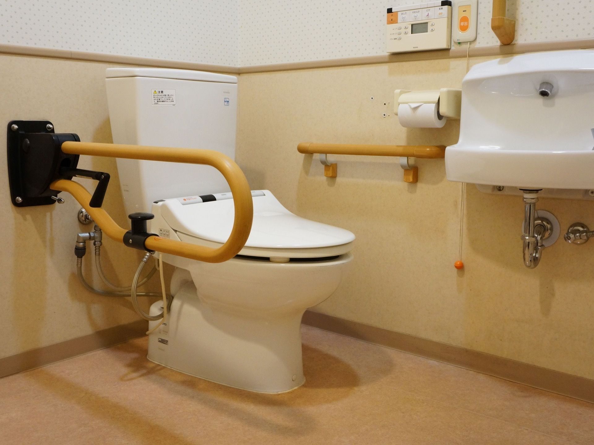 トイレの両側には、ベージュ色の大きな手すりが取り付けられている。ペーパーホルダーの右横には、小型の手洗い器もある。