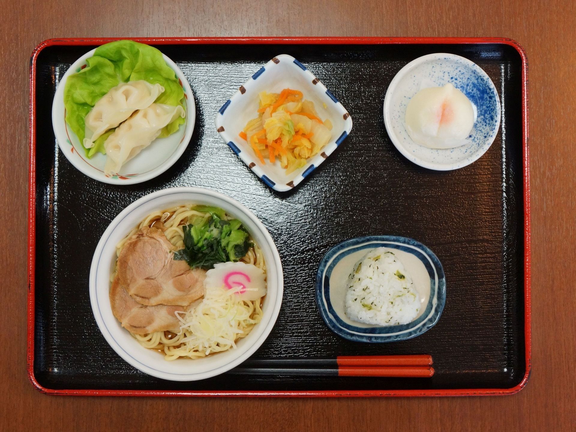 黒いトレーの上に、チャーシュー麵、三角おむすび、桃まんじゅう、ギョウザ、小鉢に入ったハクサイの和え物が並んでいる。