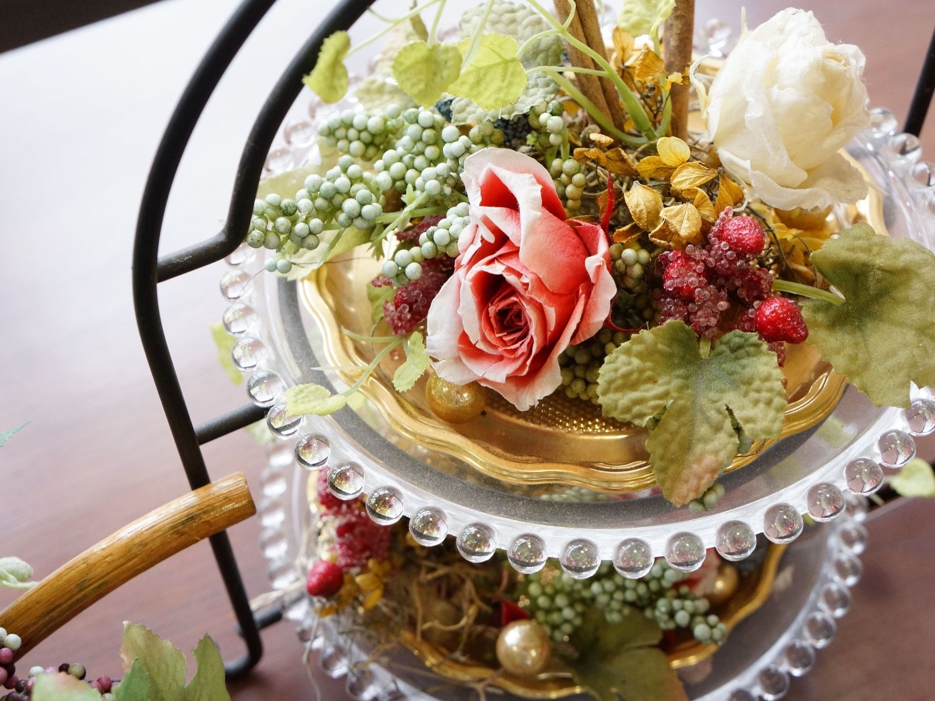 円形のガラスのケーキスタンドに飾られた、造花のフラワーアレンジメントで、ピンクと白のバラが特徴的である。