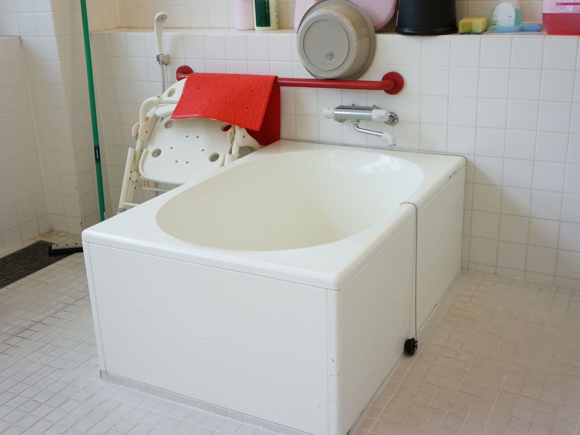 白いタイルの浴室には、浴室用のマットや椅子が用意されている。浴槽近くの壁には、赤い手すりが設置されている。