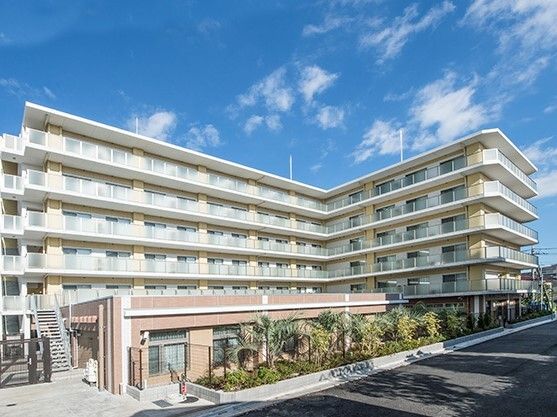 屋外階段や各フロアにベランダが付いたベージュ系の外壁で敷地内には草木が植えられている。