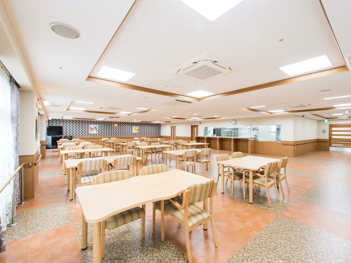 たくさんのダイニングテーブルセットが置かれた明るい雰囲気の食堂。厨房がすぐそばにある。