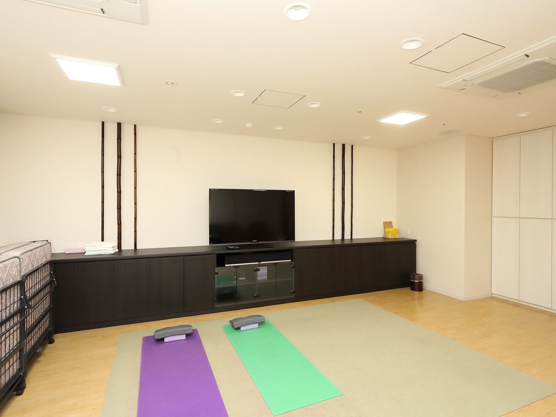 白色と木目調で構成された共有スペースである。奥には黒色の棚や大きなテレビ、折りたたみ式マットなどが置かれている。