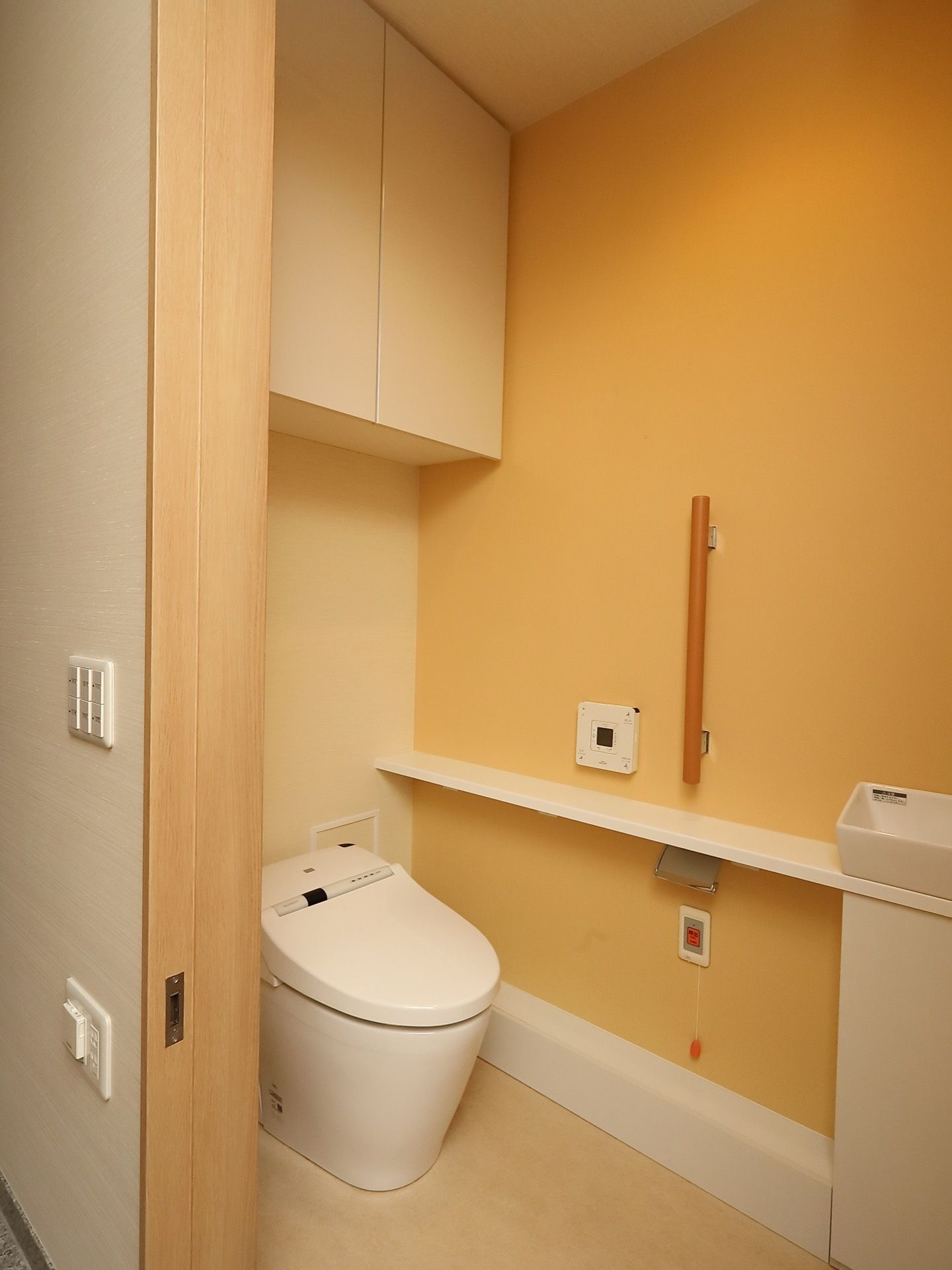オレンジ色の壁にクリーム色の床というデザインのトイレである。温水洗浄便座仕様で、壁際には緊急用ボタンや手すりも見られる。