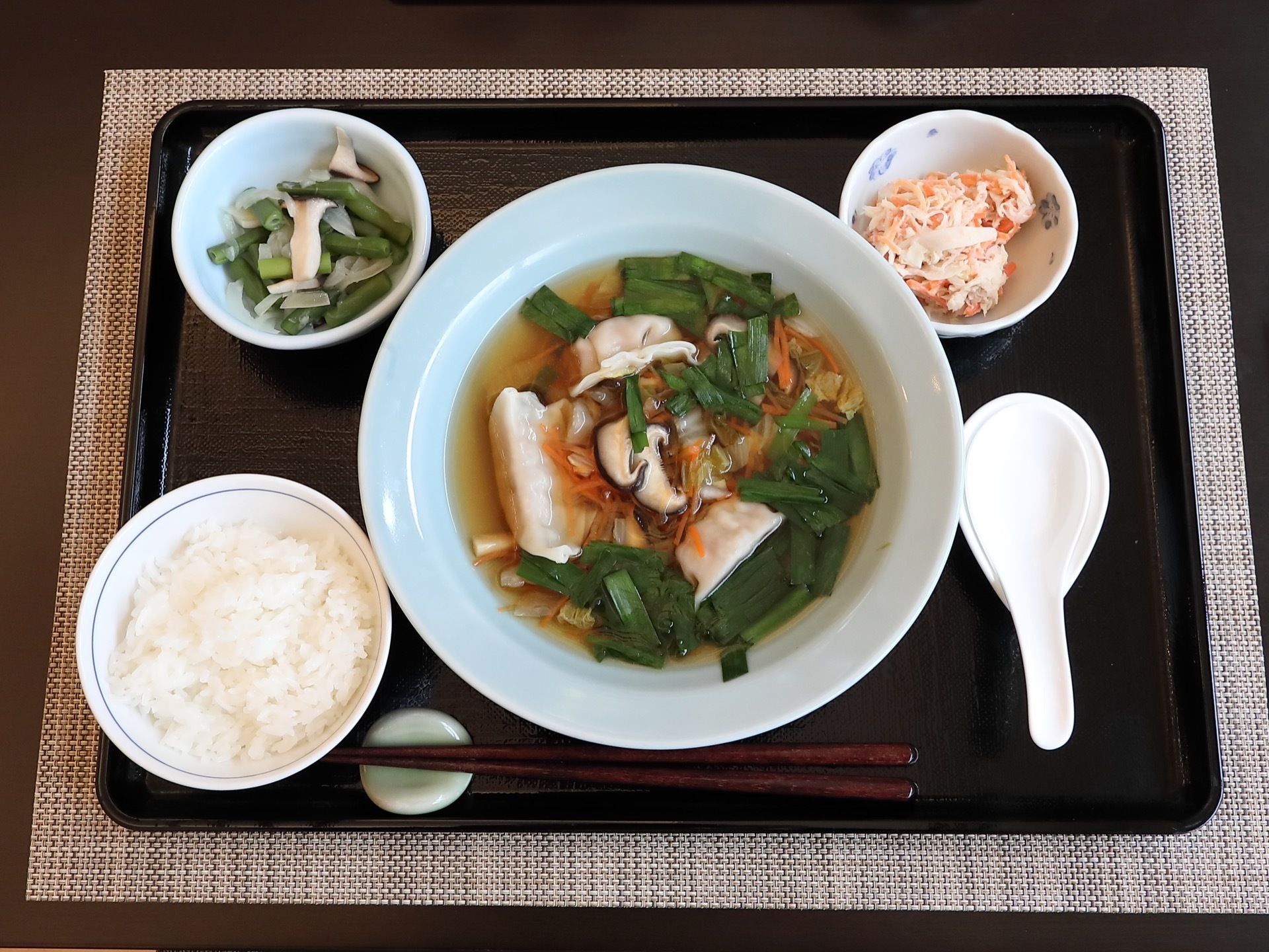 当施設で提供される食事である。黒色のお盆にご飯とメインのおかず、副菜が盛られた茶碗や皿、箸が置かれている。