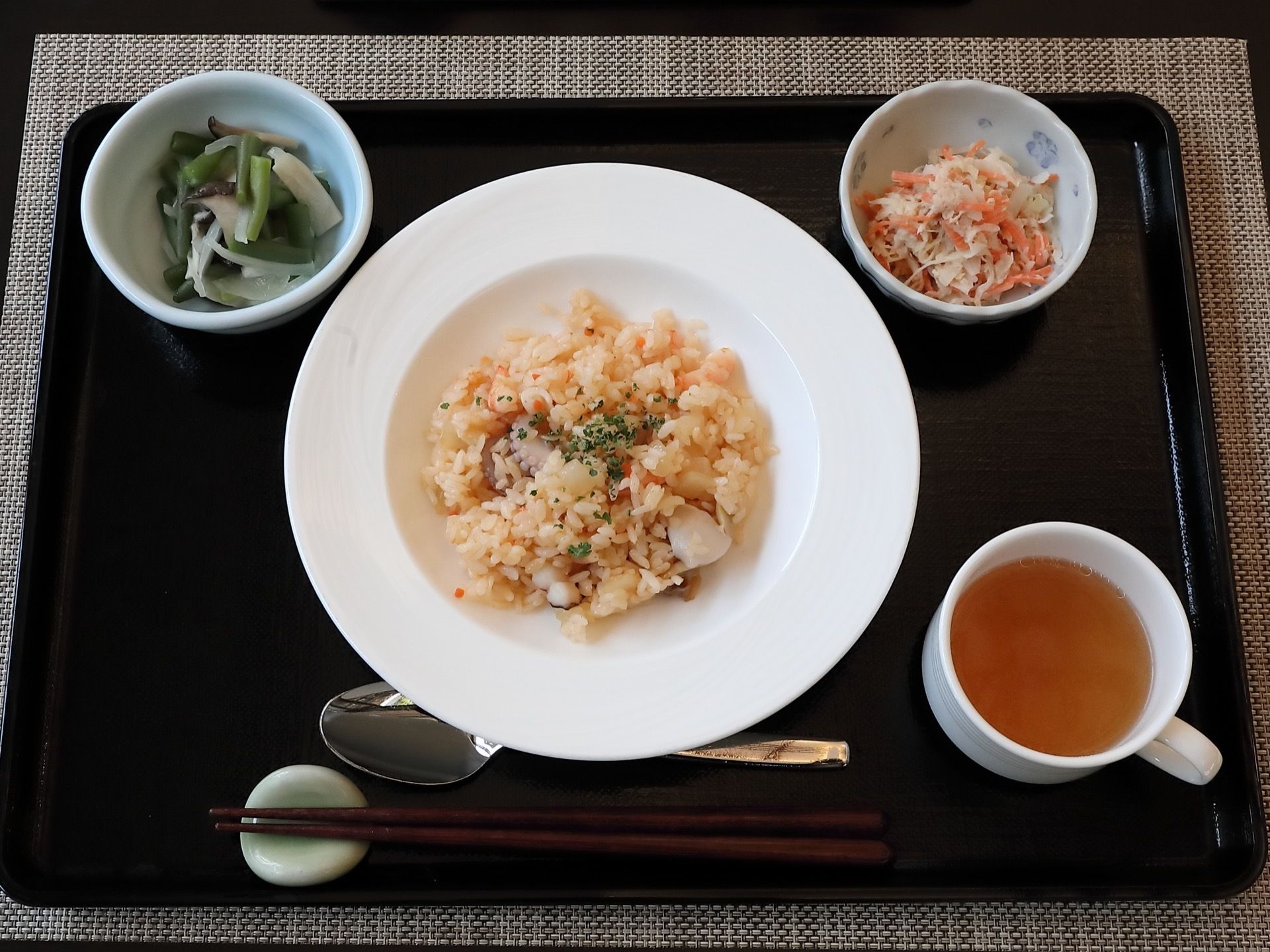 施設で提供される食事の一例である。黒色のお盆にご飯やおかず、スープが盛られた茶碗や皿、箸、スプーンが置かれている。