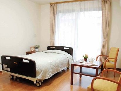 寝具がセットされたベッドが置かれたフローリングの居室。大きな窓にカーテンが付き、棚やテーブルの上には花も飾られている。