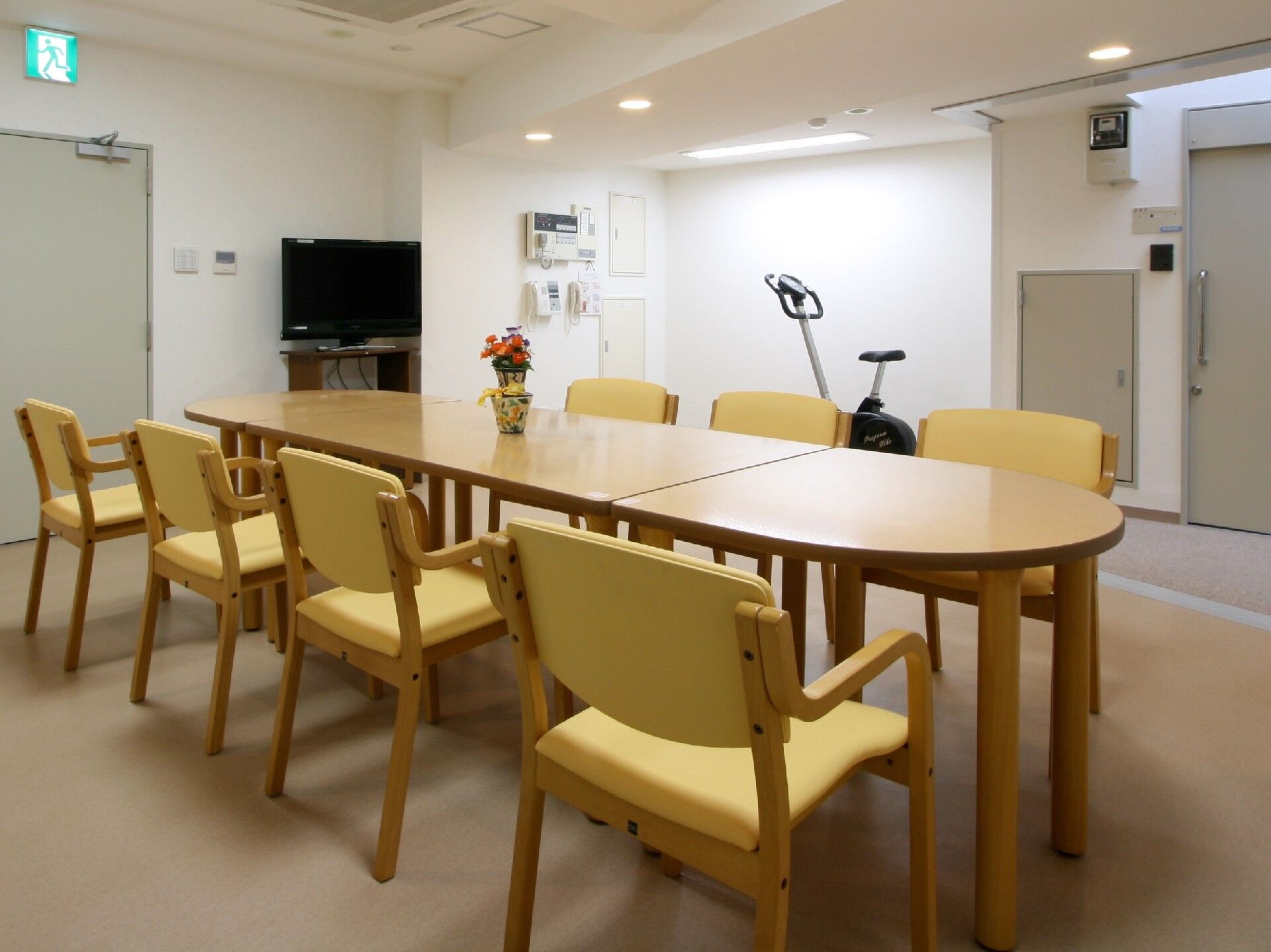 黄色い椅子がセットされた木製のテー部には花が飾られ、奥にはテレビやリハビリマシンも置かれている。