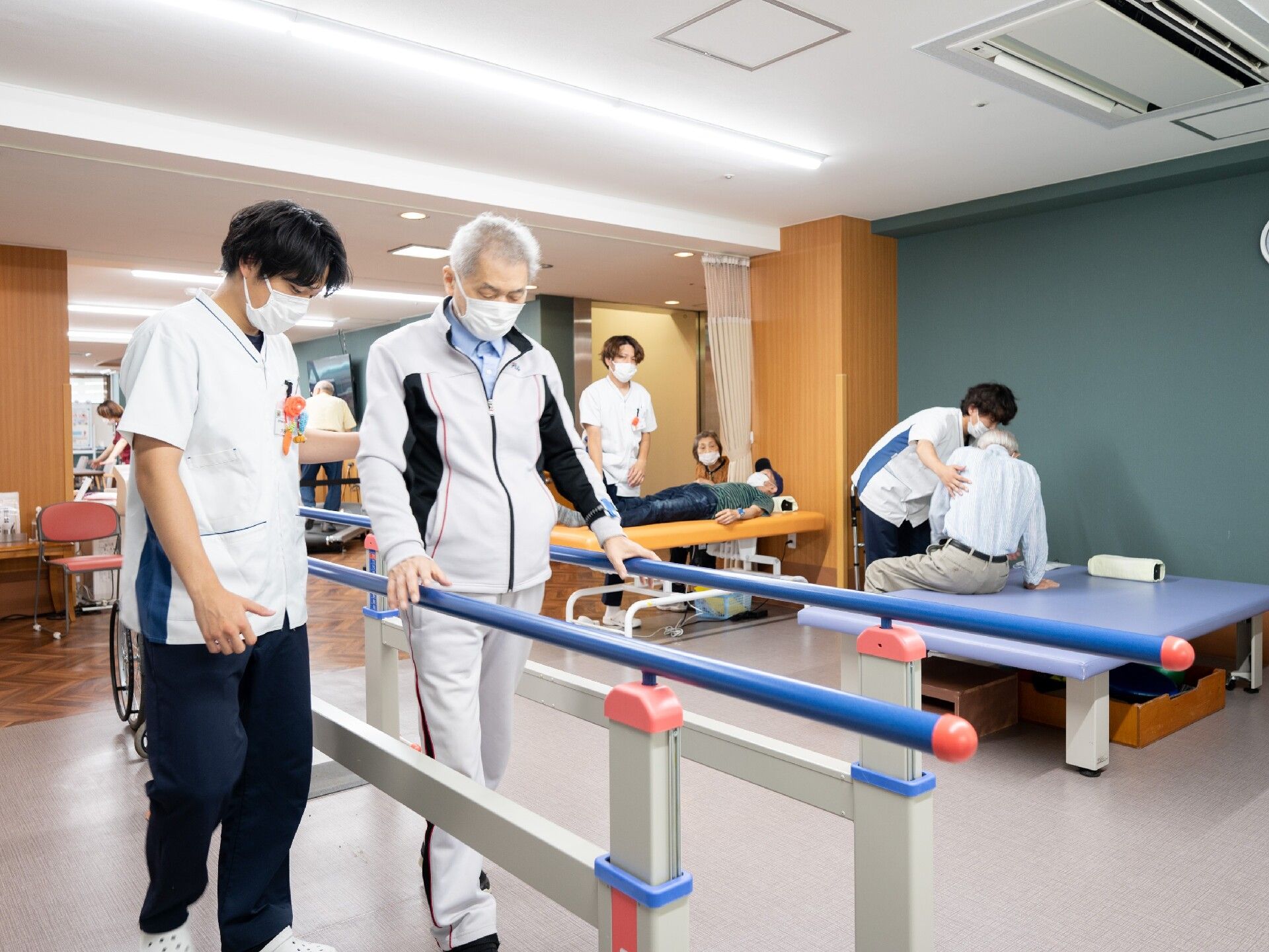 リハビリスタッフが平行棒を使って歩行練習をしている男性や、ベッドに座っている男性のサポートに当たっている。