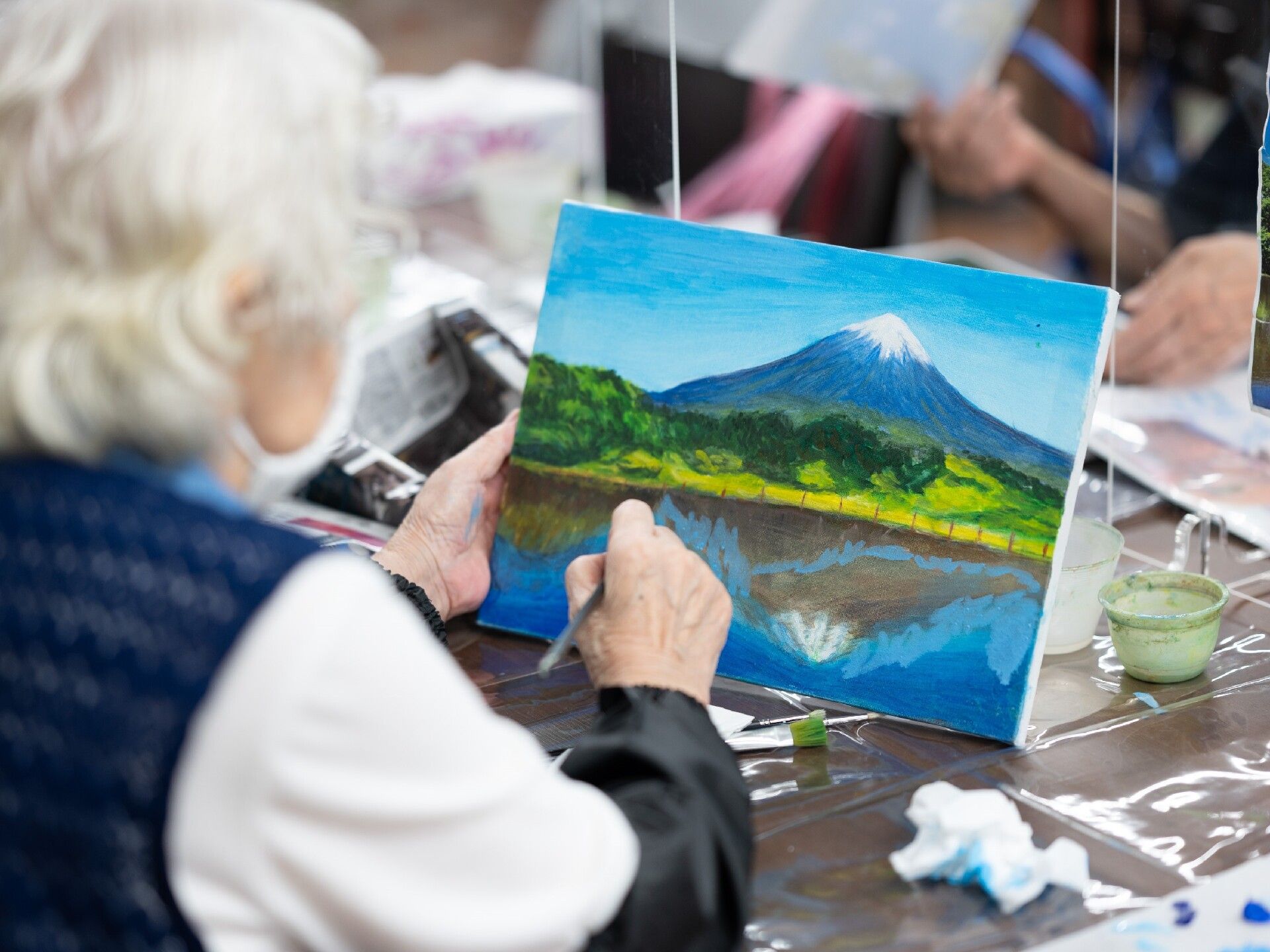 富士山の絵を絵の具で描いている白髪の高齢女性。右手には筆を持ち、左手にはキャンバスを持ち、テーブルの上にはシートが敷かれている。
