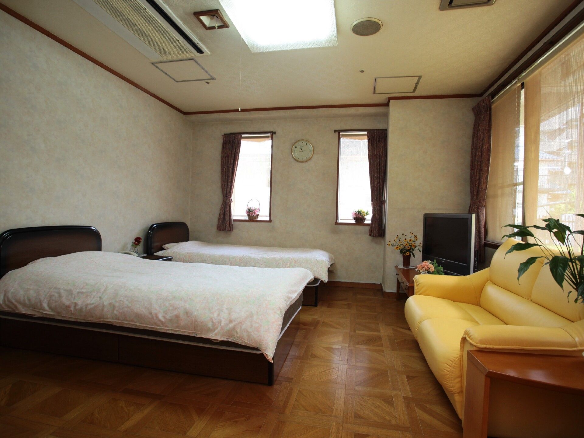 寝具がセットされたベッドが二つ置かれ、大きな窓が付いたフローリングの部屋には、黄色いソファやテレビもある。
