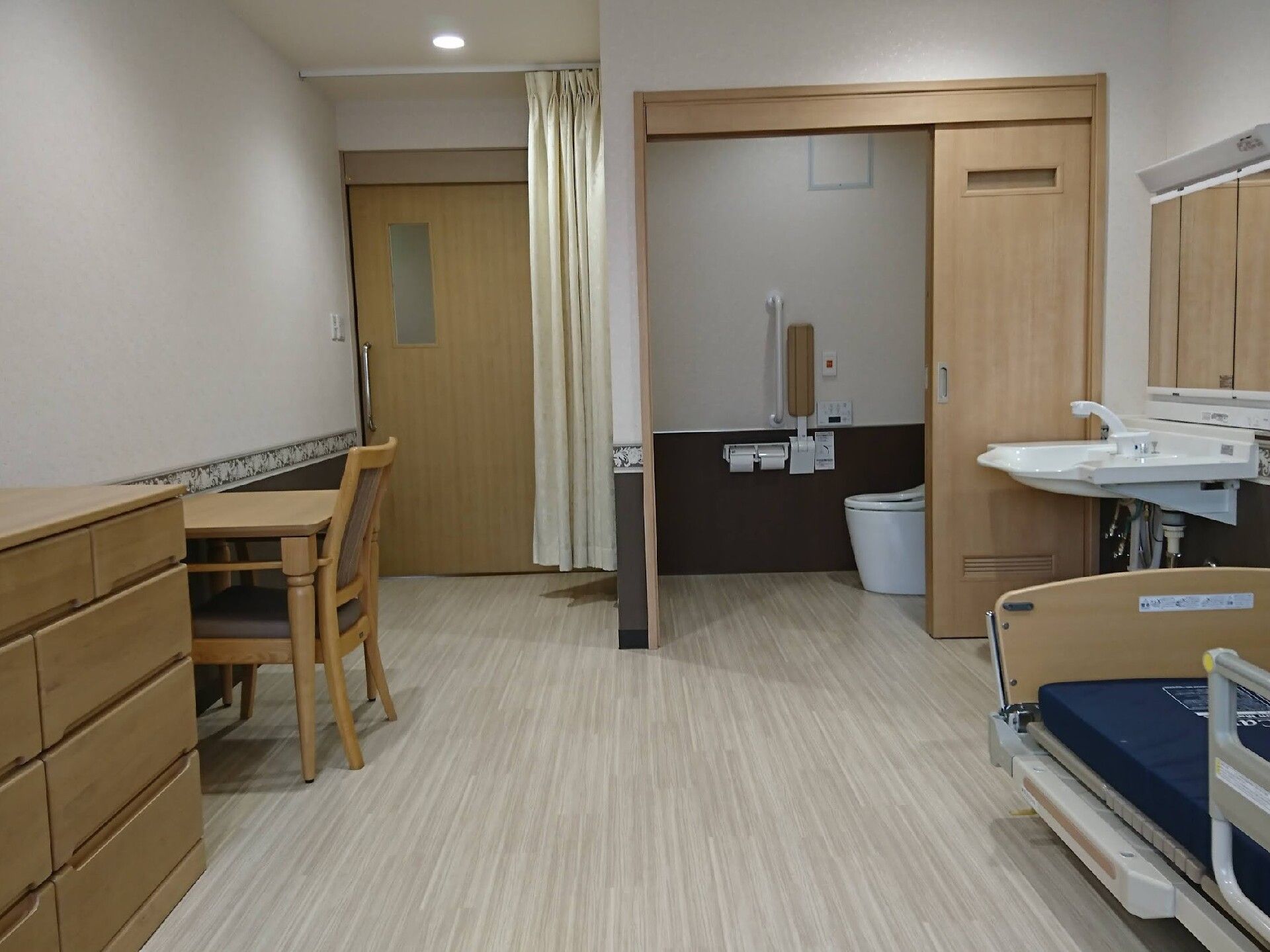 介護ベッドやタンス、テーブルセットが置かれたフローリングの部屋にはトイレも付き、居室のドアのそばにはカーテンもある。