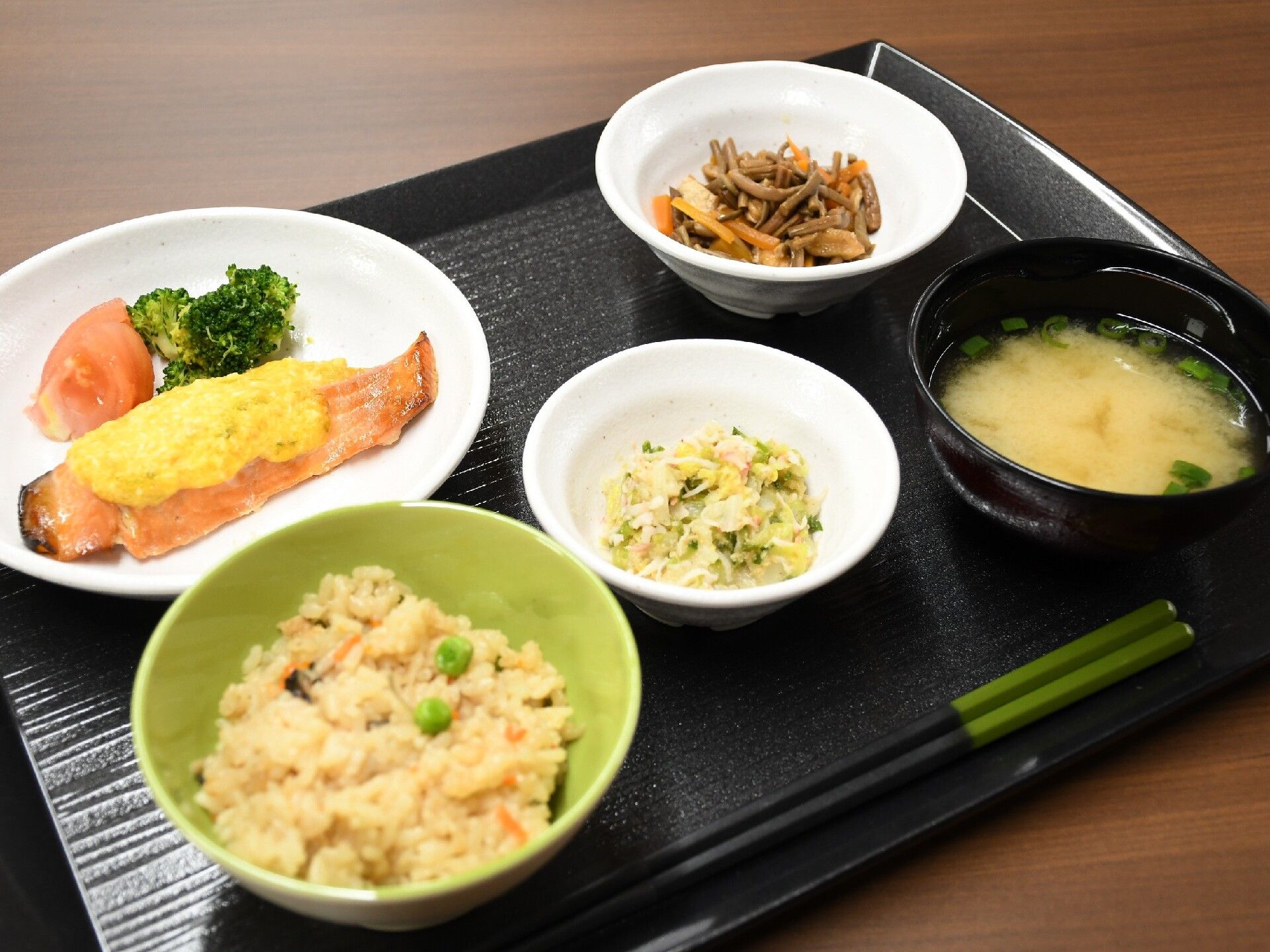 炊き込みご飯やみそ汁、野菜の和え物、鮭のソテーなどがそれぞれの器に盛られ、黒いトレーに並べられている。